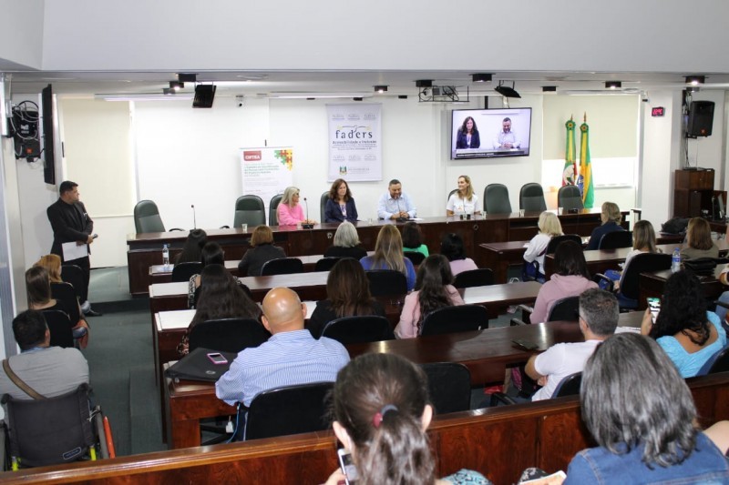 Um homem e três mulheres estão sentados atrás de uma mesa. Atrás deles, banners da Ciptea e da FADERS, uma televisão que mostra a transmissão ao vivo do evento, e as bandeiras do Rio Grande do Sul e do Brasil. À frente deles, o público, com a maior parte deles sentados atrás de mesas distribuídas pelo Plenarinho da Assembleia Legislativa.