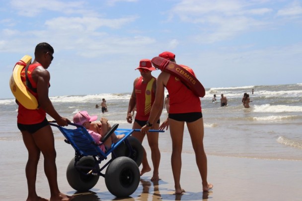 foto colorida dos guarda-vidas levando pessoa em cadeira para o banho assistido