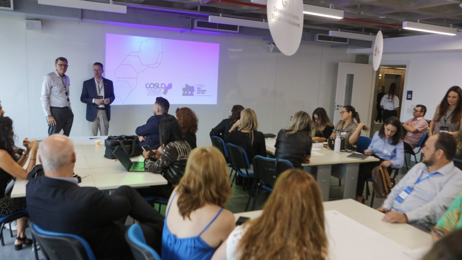 Os secretários Fantinel e Sossela estão de pé e um grupo de pessoas sentadas acompanham a apresentação do Cosud.