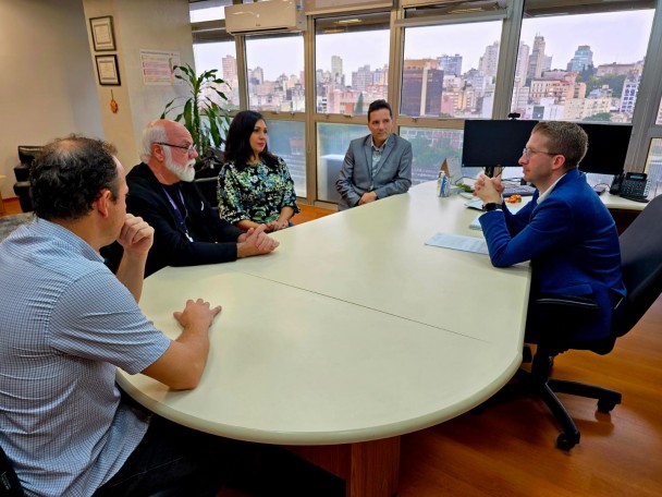 A imagem mostra cinco pessoas sentadas em volta de uma mesa branca, conversando. São quatro homens e uma mulher. Ao fundo uma janela envidraçada, através da qual se podem ver diversos prédios da cidade. A sala está situada no oitavo andar de um edifício.