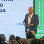 A imagem mostra um homem branco, calvo, falando ao microfone em um púlpito para uma plateia. Ele veste um terno azul e camisa branca. Ao fundo um telão com uma série de palavras e ícones, predominantemente em tons de verde.