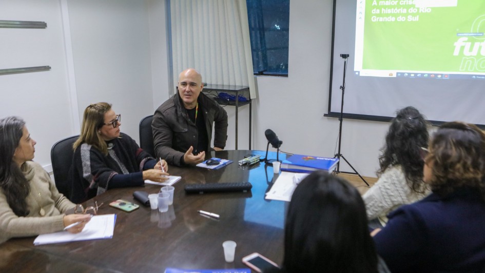 A imagem mostra seis pessoas sentadas em roda de uma mesa marrom, em reunião. A sala possui paredes brancas. Uma das pessoas, um homem branco e calvo, está com a palavra. Sobre a mesa há objetos diversos, como copos plásticos, canetas e papéis.