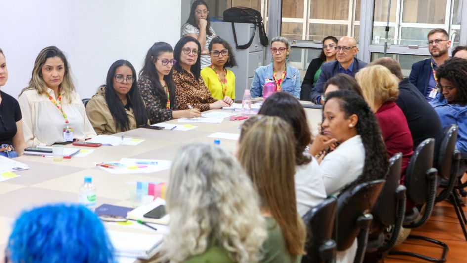 A imagem mostra cerca de 20 pessoas, predominantemente mulheres, sentadas em volta de uma mesa. A sala tem paredes brancas e uma janela ao fundo da imagem. Há diversos papéis sobre a mesa.