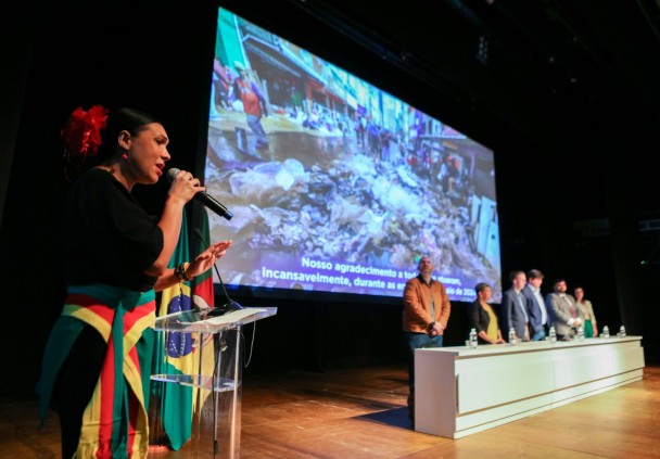 a servidora vestida com as cores da bandeira do Rio Grande do Sul com o microfone em mãos, no painel passam imagens da enchente